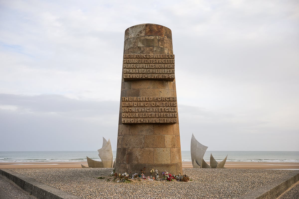 Tall stone memorial monument with engraved inscriptions and steel sculptures standing on the shore of Omaha Beach in Normandy, France, commemorating the Allied D-Day landings of 1944.
