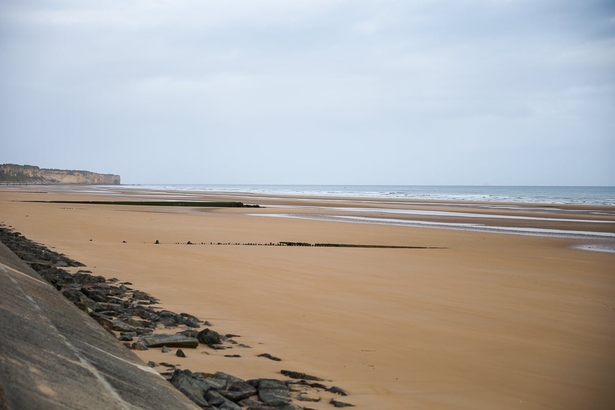 Wide view of Omaha Beach in Normandy, France, showing the expansive sandy shoreline, shallow waves, tidal flats, and cliffs along the coast, one of the main D-Day landing beaches.