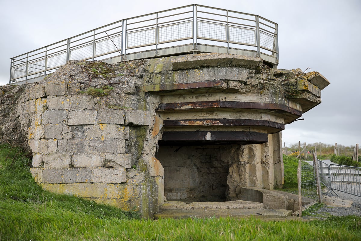 A weathered concrete WWII coastal bunker at Pointe du Hoc with heavy structural damage, located near a grassy field and protected by a metal fence.