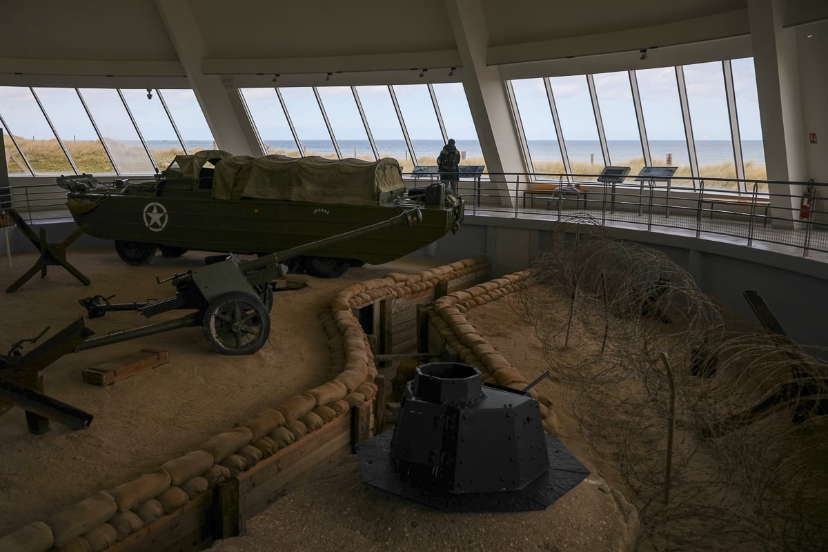 Interior of a D-Day museum featuring a DUKW amphibious vehicle, a field artillery gun, and a replica trench with barbed wire overlooking the coastline.
