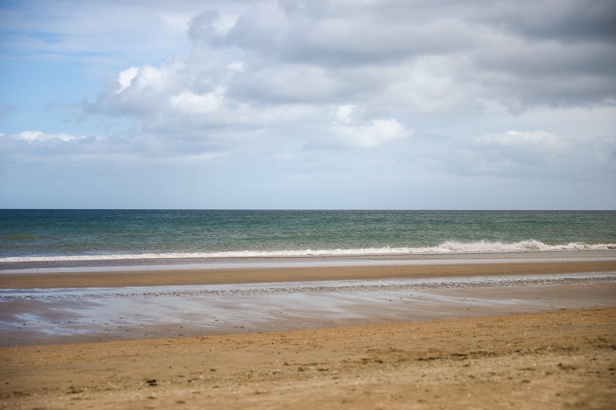 A scenic view of a quiet, sandy beach with gentle waves lapping the shore under a partly cloudy sky, representative of the D-Day landing zones.