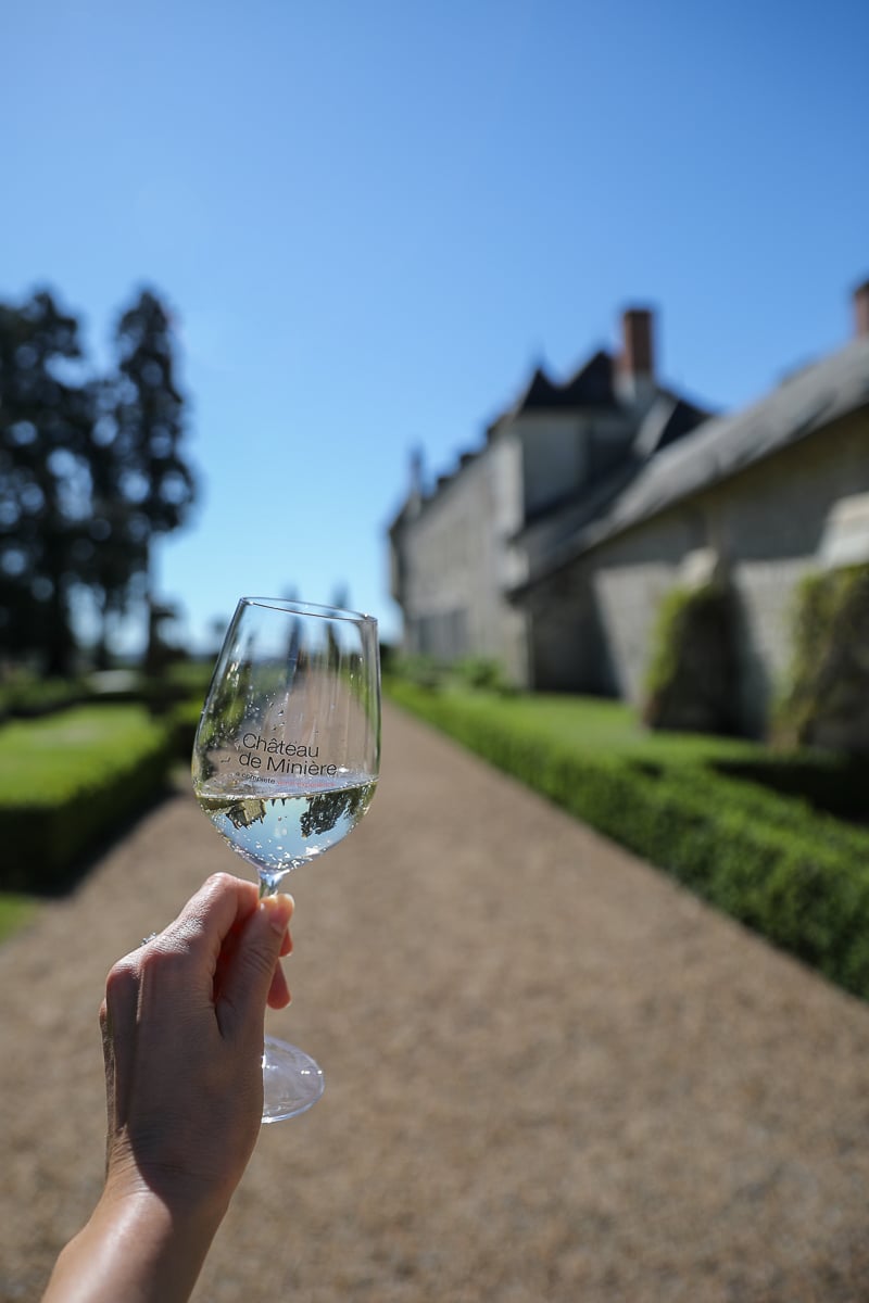A hand holds a glass of white wine in the foreground, with a historic French château and a gravel path bordered by hedges visible through the glass under a bright blue sky.
