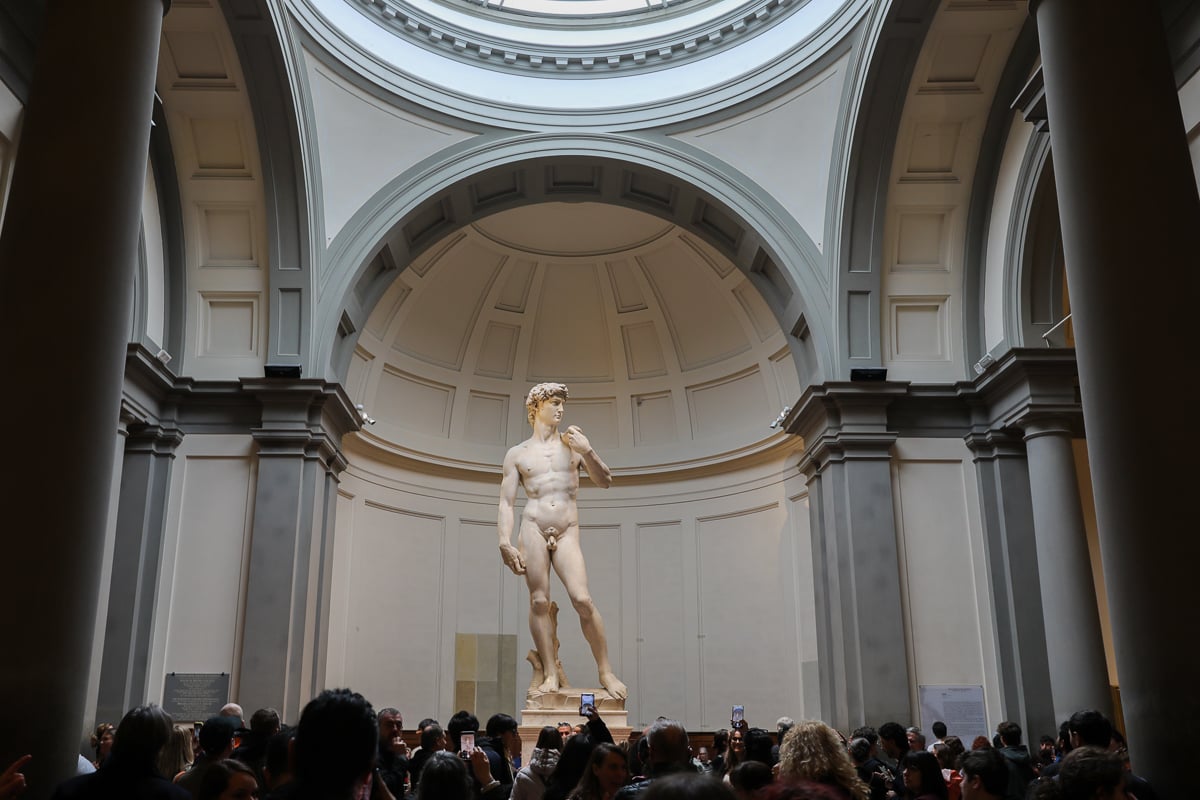 Full-length view of Michelangelo’s David, at Florence’s Accademia Gallery, beneath the domed Tribune and with crowds in front of him taking photos.