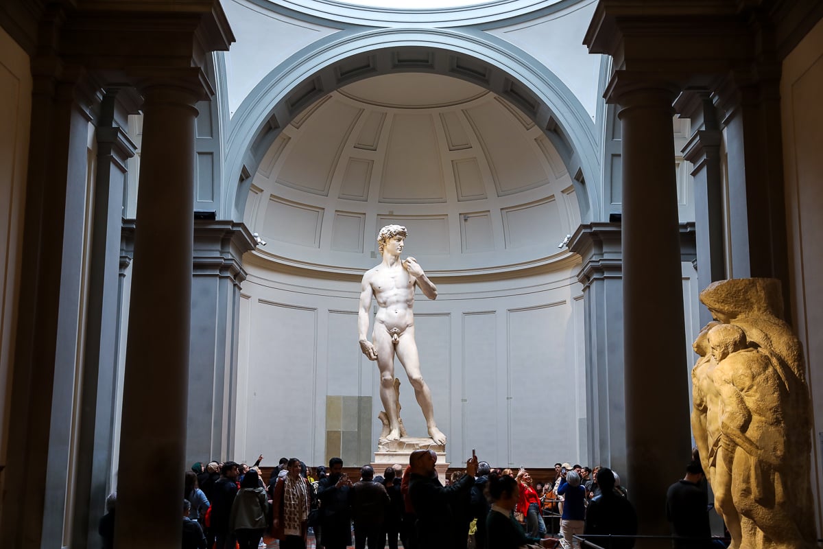 Michelangelo’s David at the end of the gallery, with an unfinished Prisoner sculpture and visitors inside Florence’s Accademia Gallery.